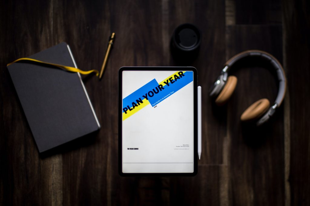 A photo with headphones, a notebook and pen, with a tablet that has the words "Plan your Year" on its screen