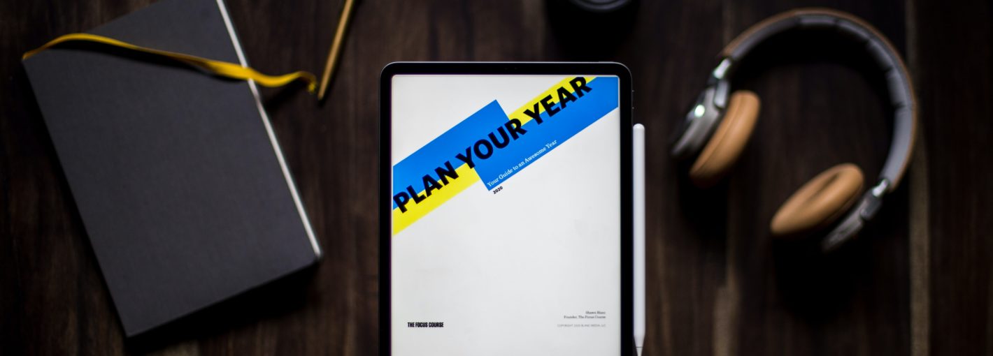 A photo with headphones, a notebook and pen, with a tablet that has the words "Plan your Year" on its screen